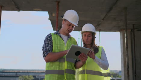 Two-engineers-a-man-and-a-woman-in-white-helmets-with-a-tablet-computer-at-the-construction-site-condemn-the-construction-plan-of-the-building